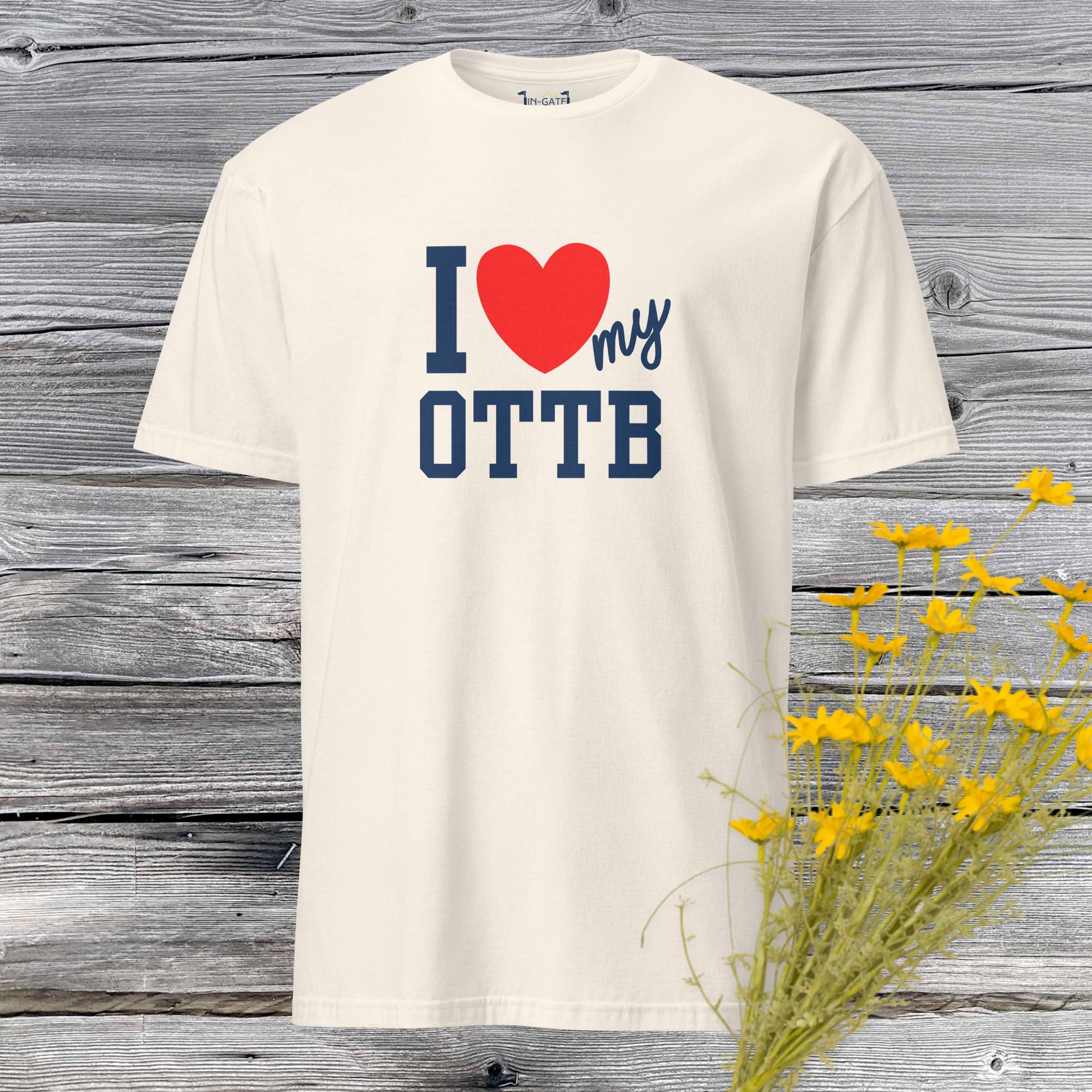 a short sleeved t-shirt in a light beige color with the I heart my OTTB logo on the front placed against a weathered barn board wall with yellow flowers in the corner
