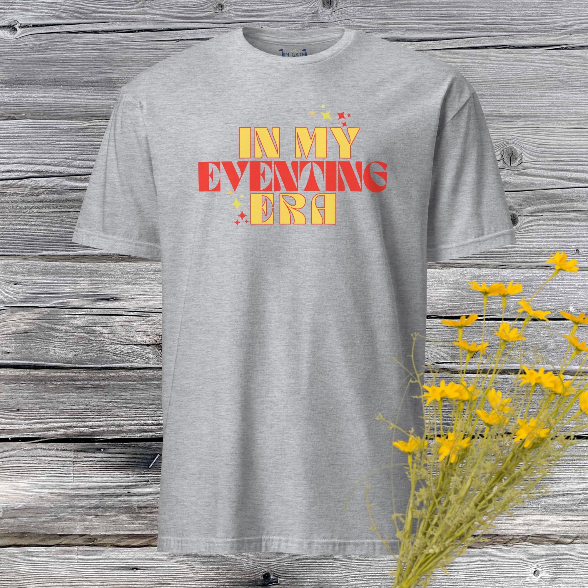 a sport gray, short sleeve t-shirt which says in my eventing era on the front in yellow and red colors against a weathered barn board wall and yellow flowers in the corner