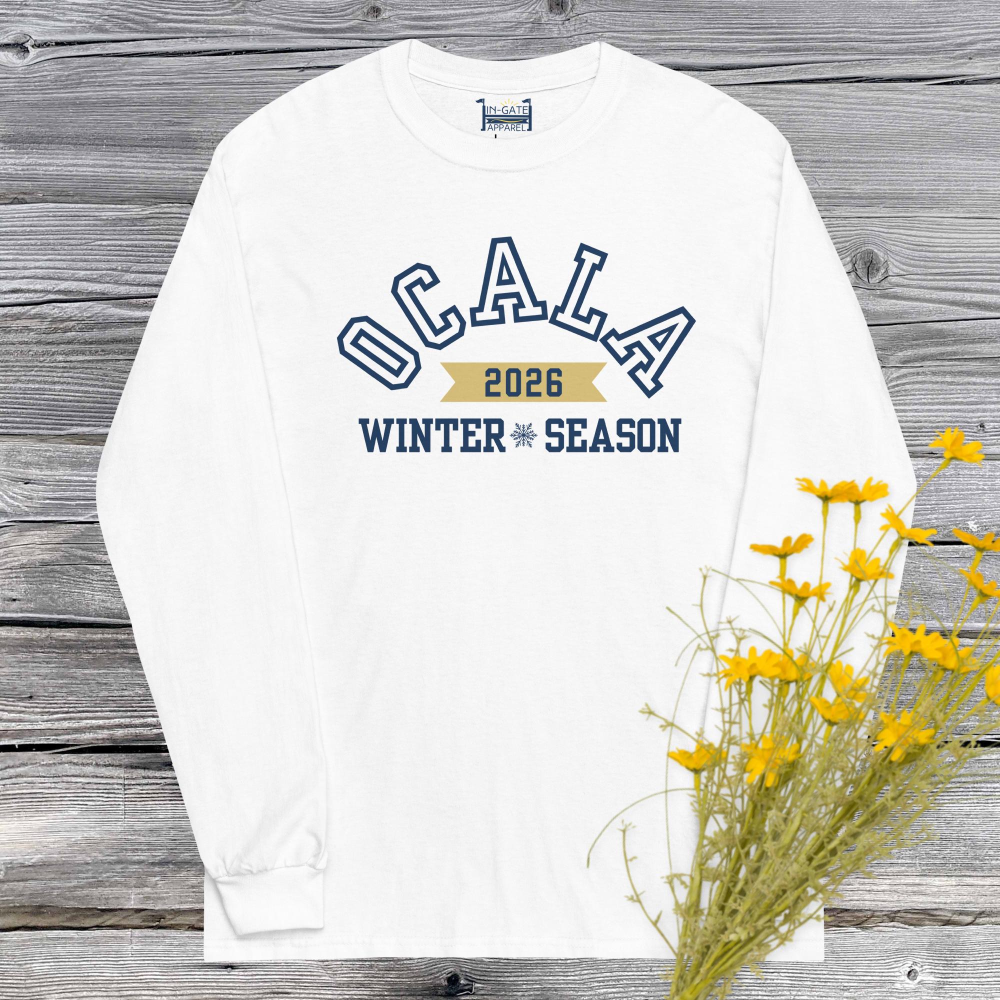 a white, long sleeve t-shirt with ocala outlined in blue arched over a gold ribbon with 2026 in it above the words winter season in solid blue hanging against a weathered barn board wall with yellow flowers in the corner