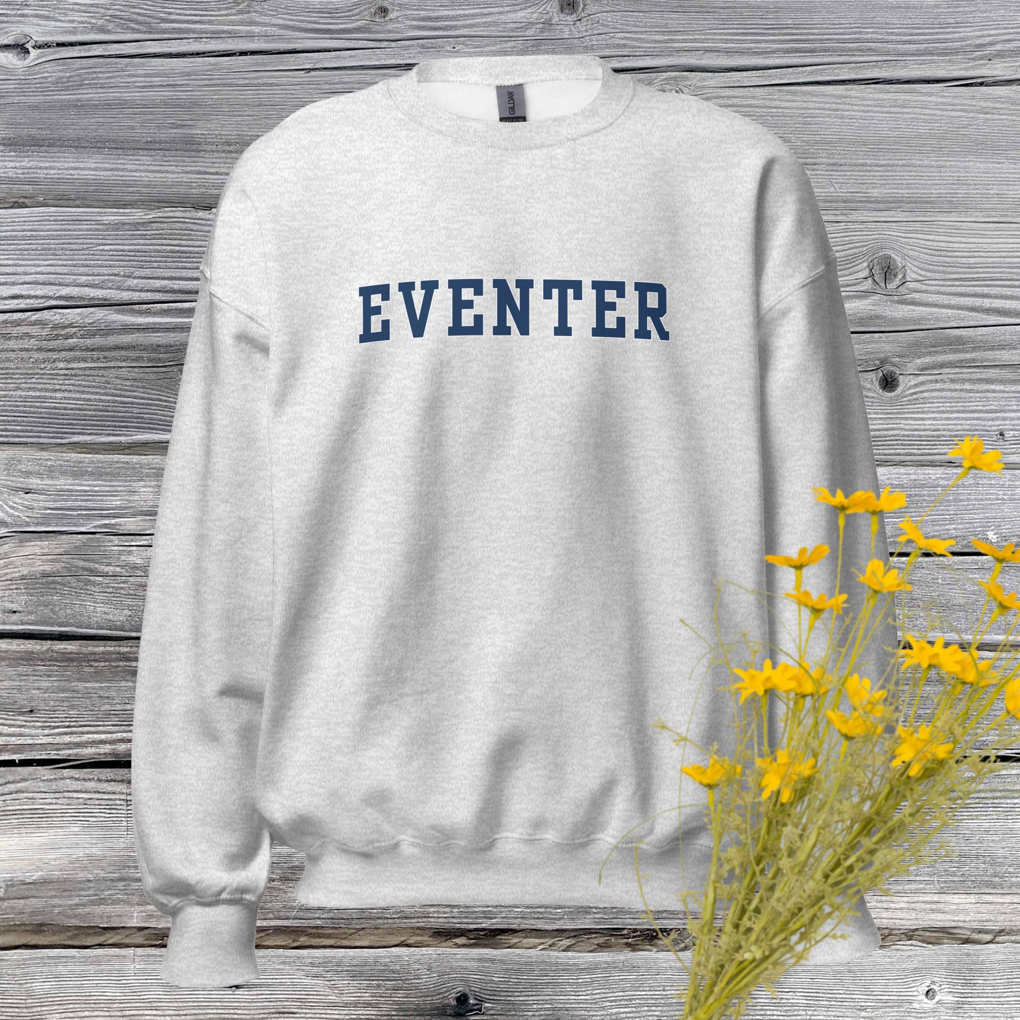 ash gray sweatshirt which says "eventer" in blue letter hanging against a weather barn board wall with yellow flowers